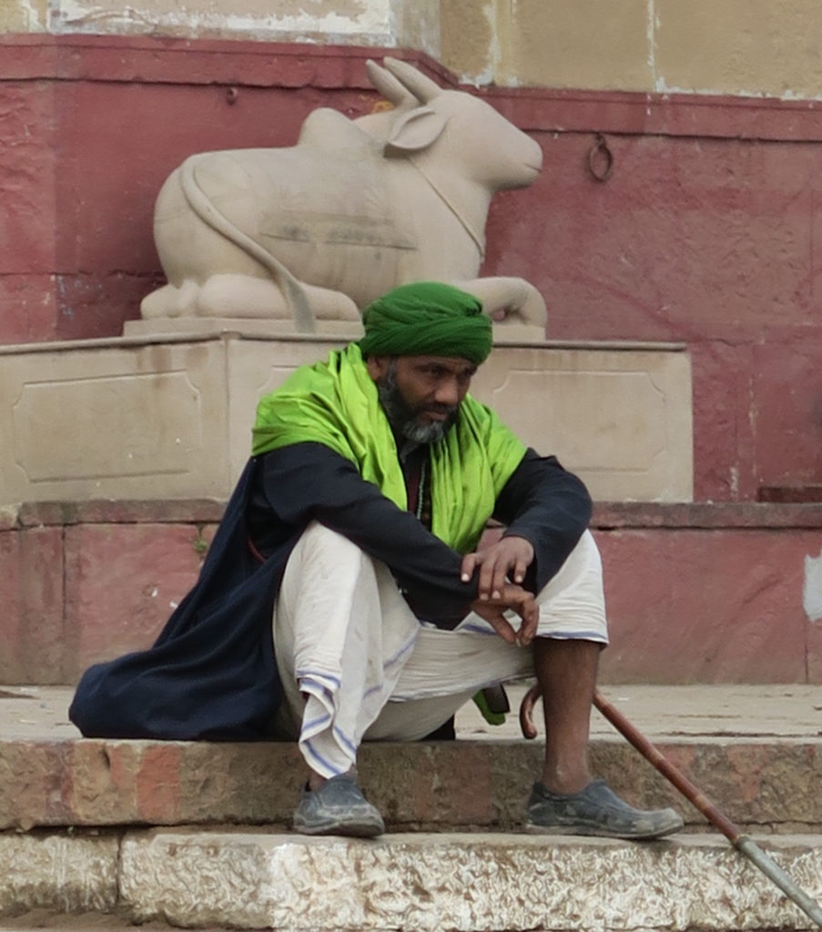 Un homme vêtu de vert avec une canne est assis devant une statue de vache